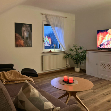Wohnzimmer der Ferienwohnung Hahnekräh: Abendstimmung mit Kerzen auf dem Couchtisch, Blick von der Couch zum Smart-TV und zum Fenster.